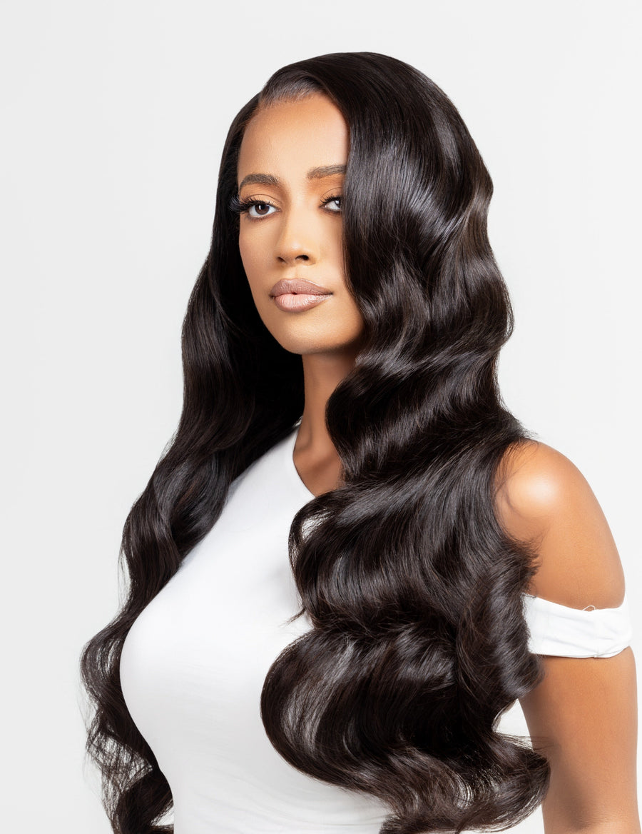 Model wearing a natural black wig with Hollywood waves, designed for alopecia, hair loss, fashion, or religious reasons. The wig features elegant, flowing curls and is styled against a white studio backdrop. The model's soft glam makeup complements the sophisticated look.